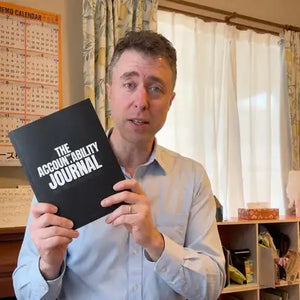 A man holding a book titled 'The Accountability Journal' in a room 